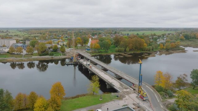 Aerial drone fly over vehicle traffic above the bridge construction site next to the forested rural area (Salacgriva city, Salaca river)