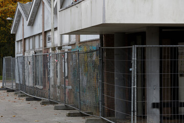 Gray metal fence surrounds a section of  graffiti-covered,  multi-story building undergoing renovation.  Autumn leaves litter the pavement.