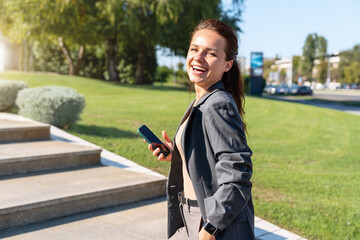 Fototapeta premium Cheerful businesswoman holding a smartphone wearing a smart casual outfit stands outdoors on a city street in summer.