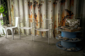 Metal room with simple interior setting containing three white metal chairs positioned in front of a weathered, rusty metal wall