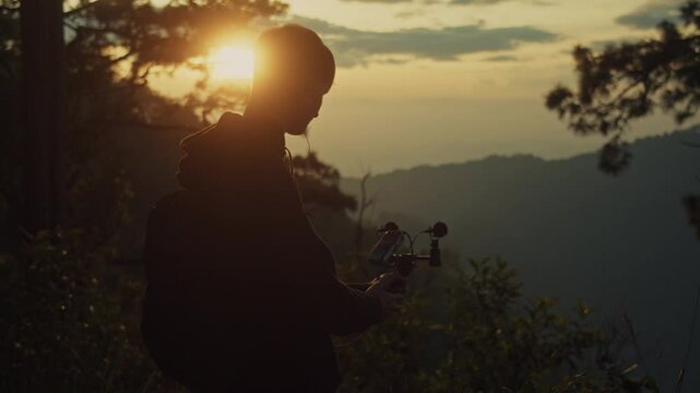 A young man holds a microphone to record his surroundings against the backdrop of a mountain sunset