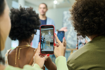 Hands of young female visitor of art exhibition or auction holding smartphone while taking photo of...