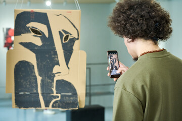 Back view of young man in t-shirt taking picture on smartphone of creative installation made up of cardboard box