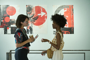 Side view of two young intercultural female visitors of modern art gallery discussing new...