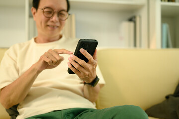 Happy elderly Asian man in eyeglasses using smartphone on couch at home