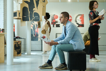 Young male visitor of modern art exhibition looking through list of expositions represented at vernissage