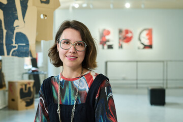 Happy young brunette female visitor of modern art gallery enjoying visiting vernissage and looking at camera
