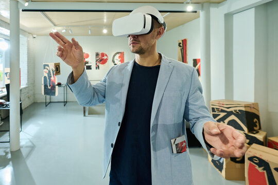 Young man in virtual reality helmet scrolling through extended artworks while standing in gallery of modern arts
