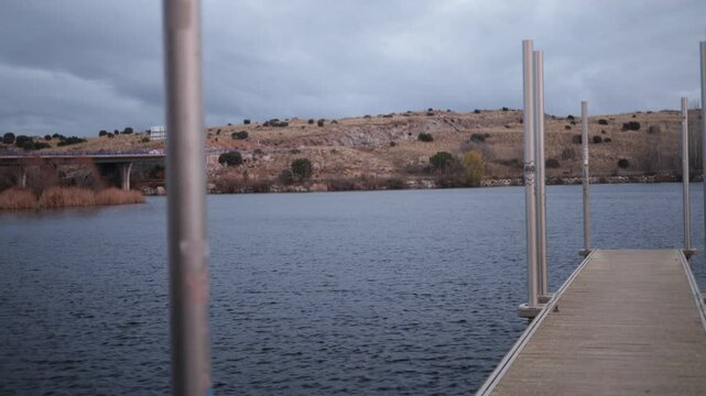 tranquil scene at Fuentes Claras in Avila, featuring a wooden dock extending into a calm lake. The overcast sky and barren hills in the background create a peaceful and reflective atmosphere
