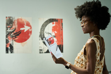 Side view of young serious African American female visitor of modern art gallery looking through brochure