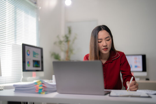 Young focused asian businesswoman in red blouse sitting at her desk taking notes from laptop with financial charts showing on computer screen in background