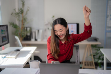 Asian businesswoman is raising her arm in celebration while looking at her smartphone and laptop,...
