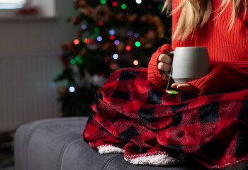 Close up of a woman holding a mug with hot drink and sitting on a sofa with a red blanket