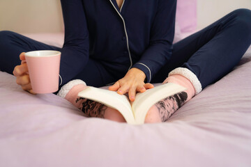 Close up shot of girl reading book with hot cup in bed in winter with pink socks