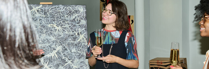 Header of young intercultural female visitors with flutes of champagne in hands listening to speaker presenting new artwork
