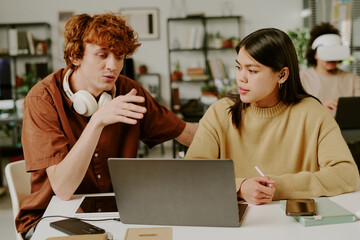 Medium close up of two IT specialists sitting at table, red haired young adult man gesticulating...