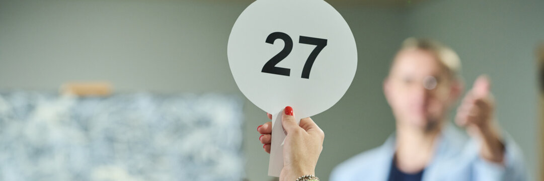 Banner of hand of young female participant of auction raising paddle with number twenty seven in front of camera