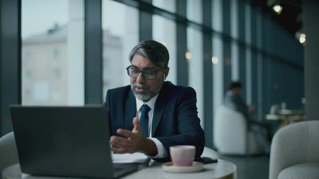 Senior Indian businessman in wireless earphones, dressed in formal attire, sitting at cafe table with coffee cup on it and having discussion via video call on laptop