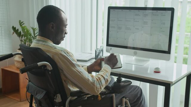 Medium long shot of African American businessman in smart casualwear using wheelchair checking his schedule on monitor at office desk
