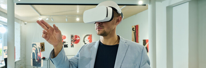 Banner of young man in virtual reality helmet scrolling through extended artworks while standing in gallery of modern arts