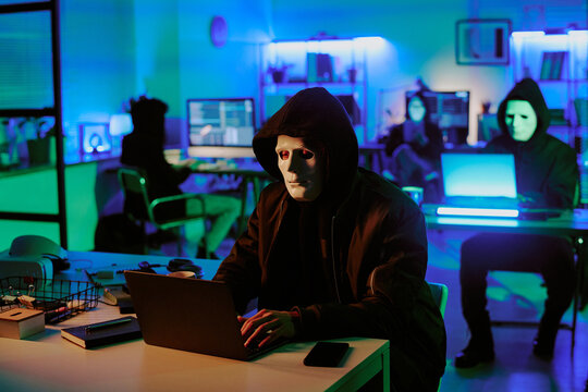 Medium close up of anonymous man in mask sitting at desk with laptop on top and coding in dimly lit room with digital devices