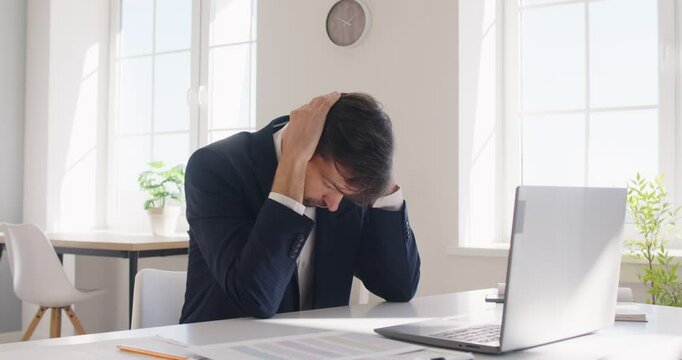 Nervous stressed young bearded man sitting at desk, talking by mobile phone in office. Unhappy frustrated guy having work problem or conflict with colleague or client, experiencing emotional distress.