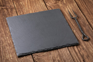 Black stone slate stand and meat fork on a wooden table, food background, template for designer