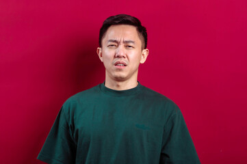 Fototapeta premium Asian man in a green T-shirt displaying a confused and slightly disgusted expression against a vibrant red background, conveying uncertainty, disapproval, or perplexity in a relatable manner