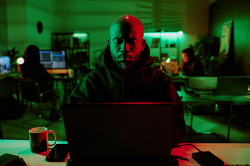 Medium close up of African American brutal hacker programming laptop in room with other hackers in...