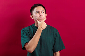 Asian man in a green T-shirt with a thoughtful expression, hand on chin, and eyes gazing upward against a vibrant red background, portraying curiosity, contemplation, or decision-making