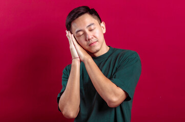 Asian man in a green T-shirt with hands resting on the side of his face and eyes closed, mimicking...