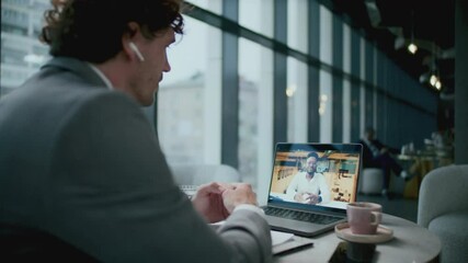 Businessman in formal suit and wireless earbuds sitting at cafe table with coffee cup and documents, discussing project with Black male colleague during video call on laptop