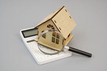 Wooden house under magnifying glass on calculator on gray background. Real estate and house price concept.