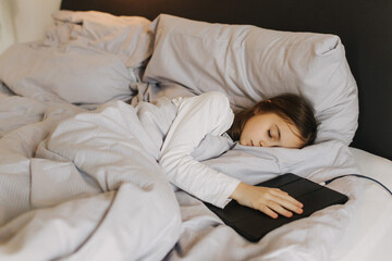 Pretty teenage girl fell asleep with a tablet in her hands, secretly watching movie, playing online games, lying under covers on bed. Generation alpha concept