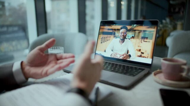 Close-up view of laptop displaying video call, young Black businessman talking to unrecognizable colleague sitting at cafe table with coffee, papers and phone