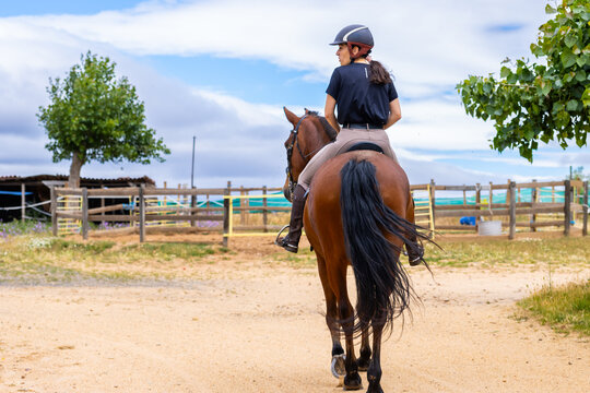 Woman riding a brown horse towards a stable on a sunny day