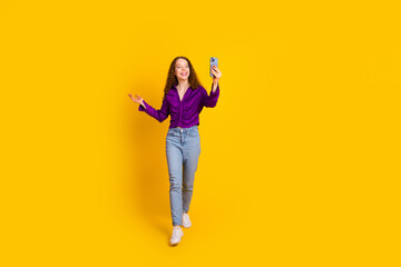 Full size photo of attractive young woman walk hold device video call dressed stylish violet clothes isolated on yellow color background