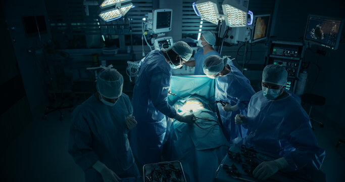 Black Male and a Caucasian Female Surgeons Performing a Hard Surgical Procedure in a Dark Operating Room Under Surgical Light. Diverse Hospital Team at Work
