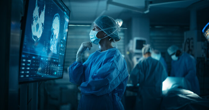 Caucasian Female Surgeon Examines Detailed Brain Scans on a Big Monitor in Operating Room. Doctor Analyzing Medical Information on Screen Before a Surgery Procedure with a Team