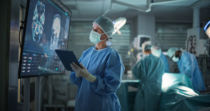 Lead Surgeon Reviews Critical Brain Scans Displayed on a Monitor as Well as Health Data on a Tablet Computer. Her Team Preparing as She Plans the Surgical Approach for the Operation