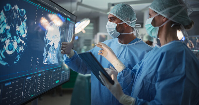 Portrait of a Young Female and Male Surgeons Reviewing Patient's Brain Scans on a Large Screen in a Dimly Lit Operating Room. Their Team of Assistants and Nurses Preparing for a Life-Saving Procedure
