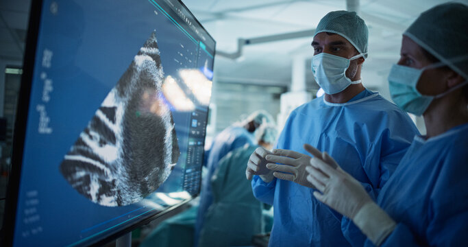 Caucasian Male and Female Surgeons Analyze Ultrasound Image on a Digital Monitor, Planning Surgical Strategy in a High-tech Operating Room. Colleagues Working Together, Preparing for Surgery