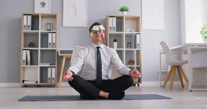 Funny man worker with post it memo work notes on face sitting in lotus position on fitness sport pilates yoga mat in business office, doing meditation breathing and calming down. Stress relief concept