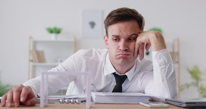 Bored exhausted company employee sitting at work desk and looking at Newton's cradle pendulum. Burnt out manager feeling unmotivated and uninspired, trying to find energy to continue his tasks.