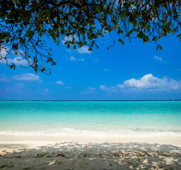 White sand beach with sea waves and splashes. Dream paradise landscape of resort for vacation and honeymoon with blue bright sky and clouds, calm turquoise lagoon, shadow of tropical tree branches