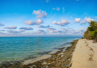 Rocky beach shore in evening, calm blue sea water with fisherman boats, sky with clouds and white sand. Tropical summer island beach in calm time of sunset, lonely silhouette of man in distance