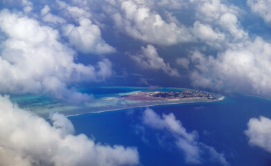 Aerial view from airplane window of Male, Maldives in scattered fluffy clouds layers. High flight of plane over blue sea and island with beach, coral reef. Scenic cloudscape and tropical climate