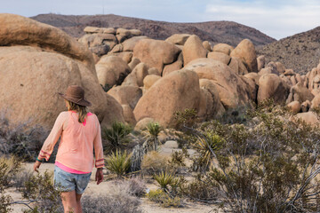 A woman with hat walking through Joshua Tree's iconic rocky terrain and desert flora under the open...