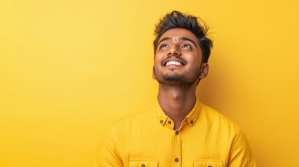 Fototapeta premium A happy young Indian man beams with a joyful smile, looking up with a sense of wonder against a vibrant yellow background. His expression reflects pure delight and enthusiasm
