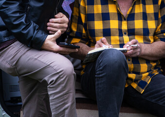 Closeup of two people collaborating outdoors, one writing in a notebook while smoking, the other holding a phone, symbolizing teamwork, creativity, storytelling and modern lifestyle.  
